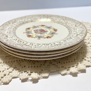 May include: Set of four white ceramic dessert plates with a gold and floral design. The plates have a scalloped edge and are stacked on top of each other.