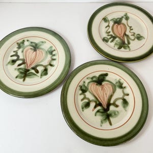 May include: Three ceramic plates with a cream-colored center, a red inner ring, and a green outer rim. Each plate features a hand-painted design of a pink heart-shaped fruit with green leaves and vines.