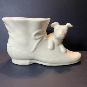 May include: A cream-colored ceramic boot-shaped planter with a dog figurine perched on the side. The boot has a bow detail and a rounded toe. The dog is lying down, adding a whimsical touch to this decorative item.