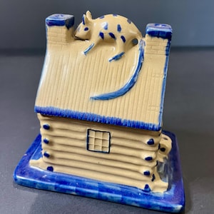 May include: A ceramic miniature cabin with a tan roof and blue accents. A blue and tan rat figurine is perched on the roof. The cabin has a small window and is set on a blue base.