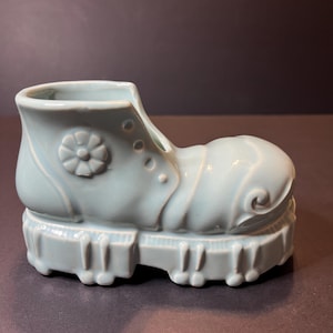 May include: A light blue ceramic boot-shaped planter. The boot features a flower design on the side, eyelets, and a ridged sole. The planter is a decorative item, suitable for small plants or as a unique home accent.