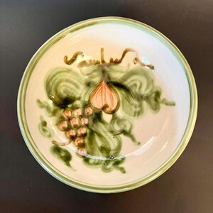 May include: A white ceramic bowl with a green rim and a hand-painted design of grapes and leaves in shades of green, brown, and pink.