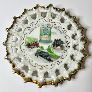 May include: A decorative white plate with a gold rim and openwork design. The center features illustrations and text for the Museum of Science & Industry Chicago, including the Mississippi, the Famous 999, and the Concord Stagecoach.
