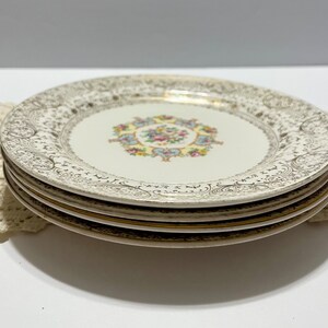 May include: A stack of four white ceramic plates with a gold rim and a floral design. The plates have a delicate, intricate pattern of gold filigree.