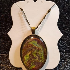 May include: Oval pendant necklace with a bronze-colored chain and setting. The pendant features a swirling abstract design in shades of brown, green, and yellow. The necklace is displayed on a white card.
