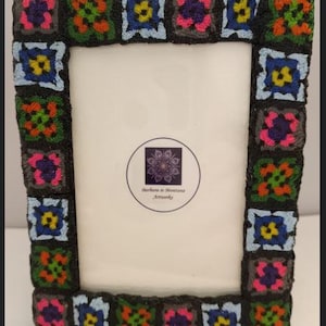 May include: A black picture frame with a colorful crocheted border. The frame is decorated with squares of different colors, including pink, blue, green, yellow, and orange. The frame is made by Barbara in Montana Artworks.