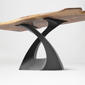 May include: A live edge wooden table with a natural, irregular edge. The tabletop is supported by a black, sculptural base with an intertwined design. The table's wood has a warm, natural color.