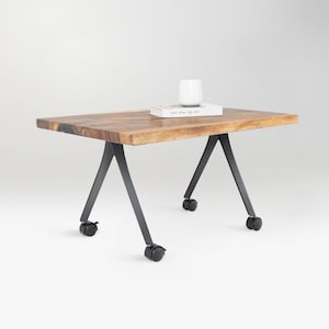 May include: A rectangular wooden coffee table with a natural wood grain finish. It has black metal legs with a unique angled design and small wheels for mobility. A white ceramic vase and a book are on top.