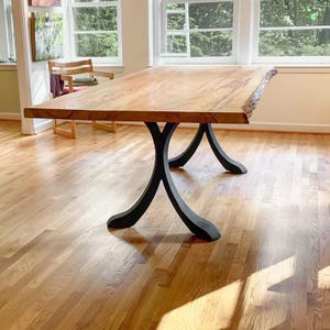 May include: A rectangular dining table featuring a natural light brown wood slab top with a live edge finish. The table is supported by two black metal legs, each designed with a curved, X-shaped base that provides structural stability.