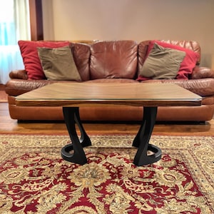 May include: A modern coffee table with a rectangular wooden top and black, curved legs. The table sits on a patterned red rug in front of a brown leather sofa with red and tan pillows. The table's dimensions are not visible.