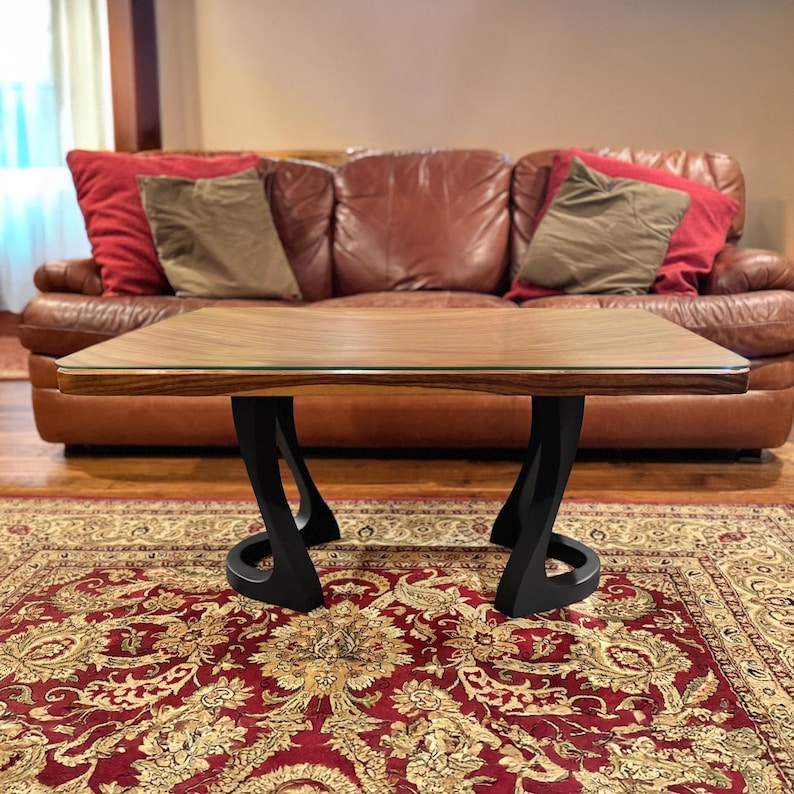 May include: A modern coffee table with a rectangular wooden top and black, curved legs. The table sits on a patterned red rug in front of a brown leather sofa with red and tan pillows. The table's dimensions are not visible.