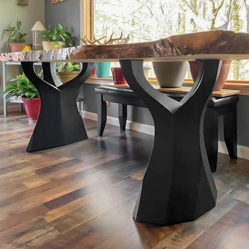 K&ouml;nnte beinhalten: Ein Esstisch aus Holz mit einer nat&uuml;rlichen, unregelm&auml;&szlig;igen Kante. Der Tisch wird von zwei schwarzen, geometrisch geformten Basen getragen. Der Tisch steht in einem Raum mit Holzb&ouml;den und einem Fenster mit Pflanzen.