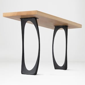 May include: A modern wooden bench with a light-colored rectangular top. The bench is supported by two sets of black, curved, oval-shaped legs. The design is minimalist and contemporary, suitable for various interior settings.