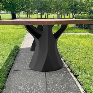 K&ouml;nnte beinhalten: Ein moderner Holztisch mit einer dunkelbraunen Tischplatte und einem schwarzen geometrischen Gestell. Der Tisch steht auf einem Betonweg mit gr&uuml;nem Gras im Hintergrund. Die Abmessungen des Tisches sind nicht sichtbar.