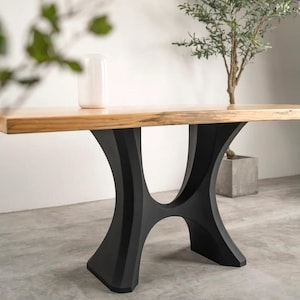 May include: A modern wooden table with a light brown top and a unique black metal base. The base has an hourglass-like design. A white vase sits on the table. The table is in a room with a concrete floor.