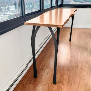 May include: A wooden console table with a natural edge and black metal legs. The tabletop is a light brown colour with a glossy finish. The legs are curved and provide a modern, artistic look. The table is placed near a window.