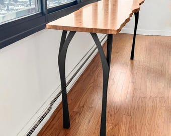 May include: A wooden console table with a natural edge and black metal legs. The tabletop is a light brown color with a glossy finish. The legs are curved and provide a modern, artistic look. The table is placed near a window.