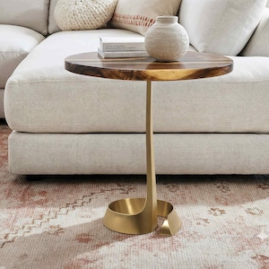 May include: A side table with a dark wood top and a gold-coloured metal base. The table is next to a cream sofa and a patterned rug. A small vase and two books are on the table. The table's height is approximately 60 cm.