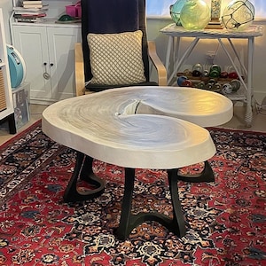 May include: A unique, light-colored coffee table with a distinctive, curved design. The tabletop appears to be made of a light-colored material with a swirling pattern. The table is supported by black, curved metal legs. The table sits on a red patterned rug.