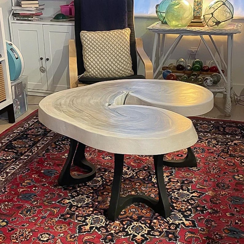 May include: A unique, light-colored coffee table with a distinctive, curved design. The tabletop appears to be made of a light-colored material with a swirling pattern. The table is supported by black, curved metal legs. The table sits on a red patterned rug.