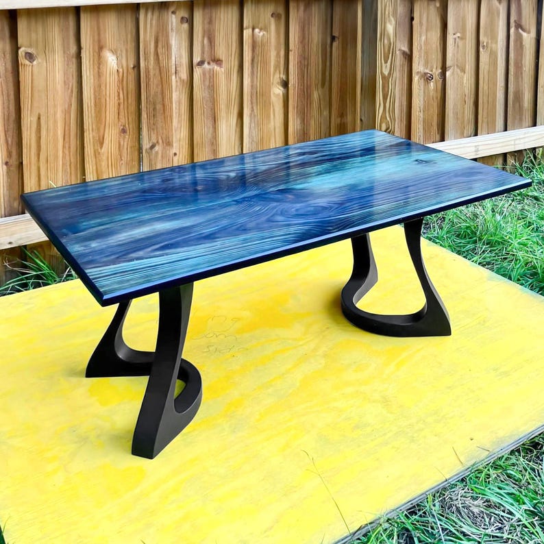 May include: A rectangular wooden coffee table with a blue and teal stained top and black curved legs. The table is set on a yellow surface, with a wooden fence and grass in the background. The table's dimensions are approximately 48 inches long and 24 inches wide.