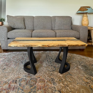 May include: A wooden coffee table with a natural edge and a black epoxy river. The table is supported by black, curved metal legs. The table is in front of a gray sofa and a patterned rug.