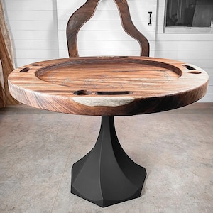 May include: A round wooden poker table with a dark, geometric base. The tabletop has carved player positions and cup holders. The wood grain is visible, and the table is set against a white door and wall.