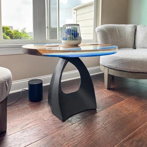 May include: A modern coffee table with a black, curved base and a wooden top with a blue epoxy resin inlay. The table is in a living room setting with a gray armchair and a window in the background.