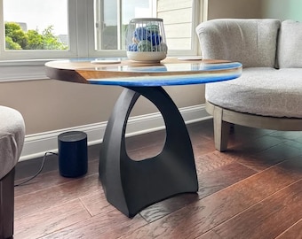 May include: A round side table featuring a natural wood grain top with a blue resin inlay along the edge. The table is supported by a solid black pedestal base with a central cutout. A white wire basket containing blue spheres sits on top.