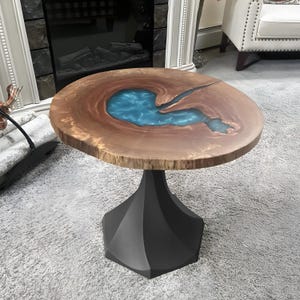 May include: A round wooden side table with a live edge and a blue resin inlay. The tabletop is supported by a black geometric base. The table measures approximately 61 cm in diameter.