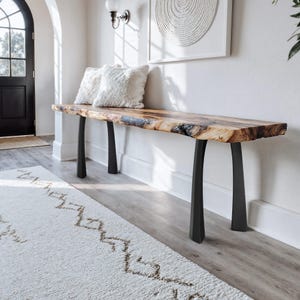 Peut inclure: Une console en bois avec un bord brut et des pieds en métal noir. La table présente un fini veiné naturel et est surmontée de deux coussins blancs moelleux. La table est placée contre un mur blanc.