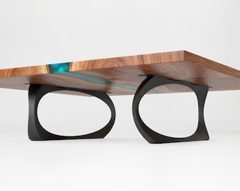 May include: A rectangular coffee table featuring a natural wood grain surface with a central translucent teal resin inlay. The table is supported by two black metal legs with a curved, open oval design attached to the underside.