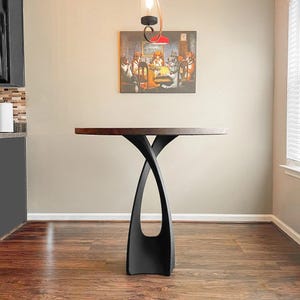 May include: A modern, dark wood-topped table with a unique, black, intertwined base. The table is set against a neutral wall with a framed painting of dogs playing poker. The room has hardwood flooring.