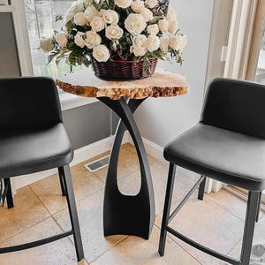 May include: A live edge wood table with a black metal base, topped with a basket of white roses. Two black bar stools flank the table. The table's top is approximately 30 inches in diameter.
