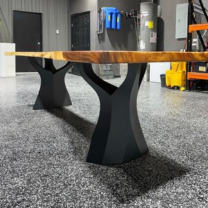 K&ouml;nnte beinhalten: Ein gro&szlig;er, rechteckiger Holztisch mit nat&uuml;rlicher Kante. Die Tischplatte hat eine warme braune Farbe und wird von zwei schwarzen, baumf&ouml;rmigen Metallf&uuml;&szlig;en getragen. Der Tisch steht in einer Garage mit einem gesprenkelten grauen Boden.