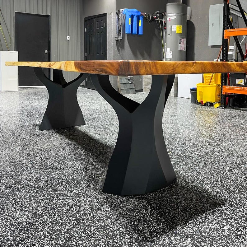 K&ouml;nnte beinhalten: Ein gro&szlig;er, rechteckiger Holztisch mit nat&uuml;rlicher Kante. Die Tischplatte hat eine warme braune Farbe und wird von zwei schwarzen, baumf&ouml;rmigen Metallf&uuml;&szlig;en getragen. Der Tisch steht in einer Garage mit einem gesprenkelten grauen Boden.
