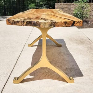 May include: A dining table featuring a natural live edge wood slab top with prominent grain patterns and dark inclusions, supported by a sculptural, Y-shaped base finished in a smooth, matte gold metallic tone with four splayed legs.