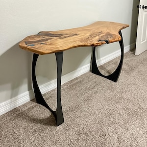 May include: A live edge console table with a natural wood top and black metal legs. The wood top has a light brown color with darker wood grain patterns and a unique, organic shape. The legs are curved and provide a modern aesthetic.