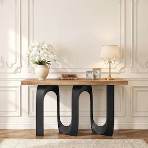 May include: A modern console table with a natural wood top and black, curved metal legs. The table is decorated with a vase of white flowers, books, framed photos, and a gold lamp. The table is set against a white wall.