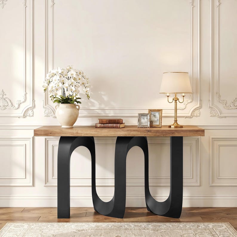May include: A modern console table with a natural wood top and black, curved metal legs. The table is decorated with a vase of white flowers, books, framed photos, and a gold lamp. The table is set against a white wall.