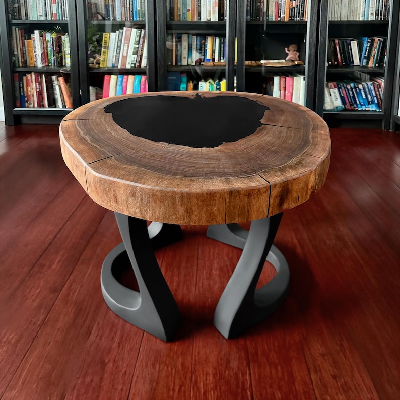 May include: A round wooden coffee table with a dark, black-filled center. The tabletop is made of a light brown wood with visible grain and cracks. The base is a dark gray, sculptural design. The table is a unique piece of furniture.