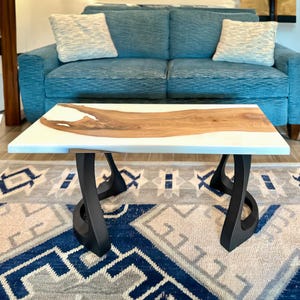 May include: A rectangular coffee table with a white surface and a natural wood inlay. The table has black, curved metal legs. The table is in front of a blue sofa with two decorative pillows. The rug has a blue and gray geometric pattern.