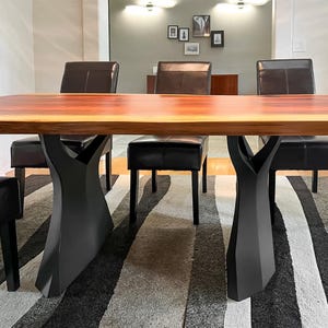 K&ouml;nnte beinhalten: Ein Esstisch mit einer rotbraunen Holzplatte und einem schwarzen, skulpturalen Untergestell. Schwarze Lederst&uuml;hle umgeben den Tisch. Der Raum hat eine neutrale Farbpalette mit einem gestreiften Teppich und gerahmten Kunstwerken an der Wand.
