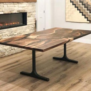 May include: A rectangular wooden dining table with a brown and tan epoxy resin top. The table has black metal legs. The table is in a room with a fireplace and a decorative wall hanging.