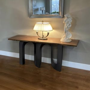 May include: A wooden console table with a live edge top and black metal legs. A lamp with a cream-colored shade and a white statue are on the table. A mirror hangs above the table.