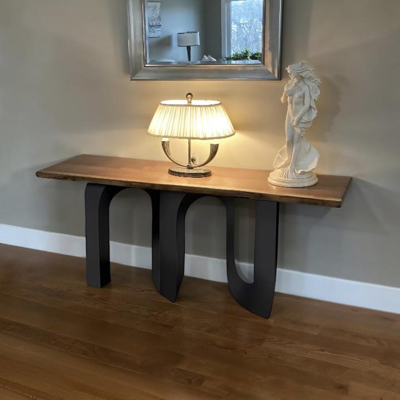 May include: A wooden console table with a live edge top and black metal legs. A lamp with a cream-colored shade and a white statue are on the table. A mirror hangs above the table.