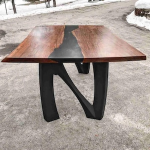 May include: A rectangular wooden table with a dark gray epoxy river running through the center. The tabletop is a rich brown color, and the legs are black and have a unique, curved design. The table is a modern piece of furniture.