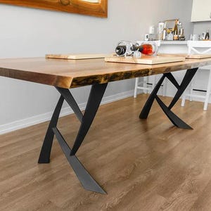 May include: A large, rectangular wooden dining table with a live edge and black metal legs. The table has a natural wood grain finish and is set on a hardwood floor. A wooden serving board and wine accessories are on the table.