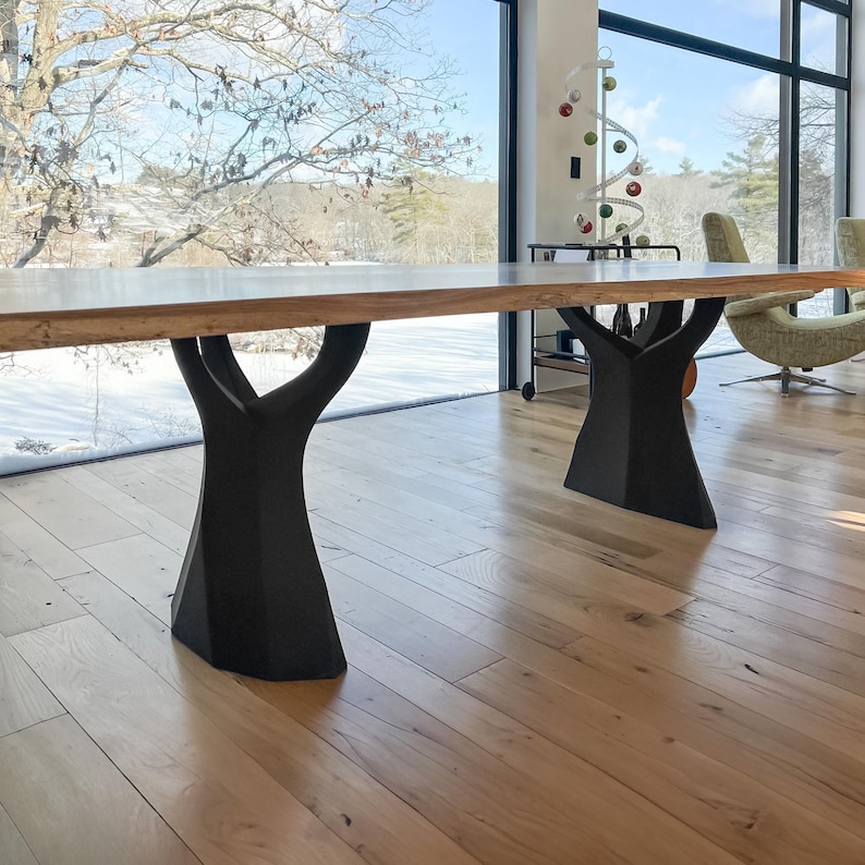K&ouml;nnte beinhalten: Ein gro&szlig;er Esstisch aus Holz mit einer hellen Tischplatte und schwarzen, baumstammf&ouml;rmigen Beinen. Der Tisch steht in einem modernen Raum mit gro&szlig;en Fenstern mit Blick auf eine verschneite Landschaft.