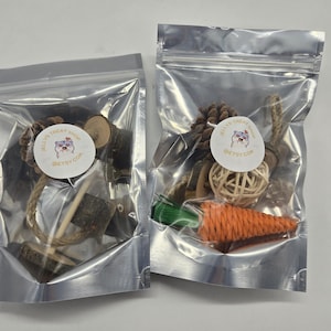 Op de afbeelding: Twee doorzichtige plastic zakken gevuld met kleine speeltjes voor huisdieren. Eén zak bevat houten blokken en touw, de andere heeft een dennenappel, een geweven bal en een wortelvormig speeltje. Elke zak heeft een ronde sticker met tekst en een cartoonafbeelding.